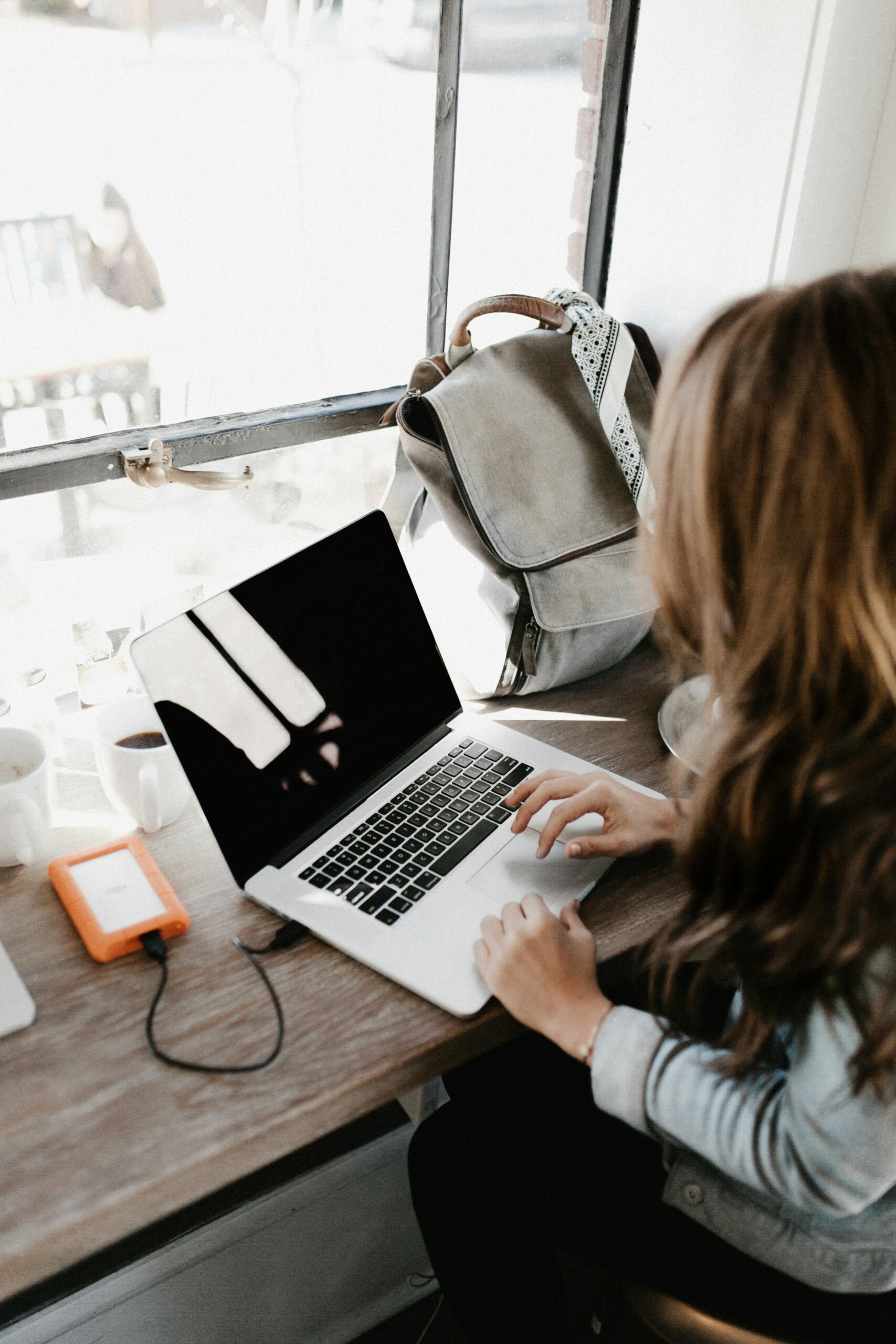 A view of a woman sitting down in a cafe on a high bench facing the window with long brown hair typing on an open laptop, with a grey rucksack to the right and a bright reflection of the window on the laptop