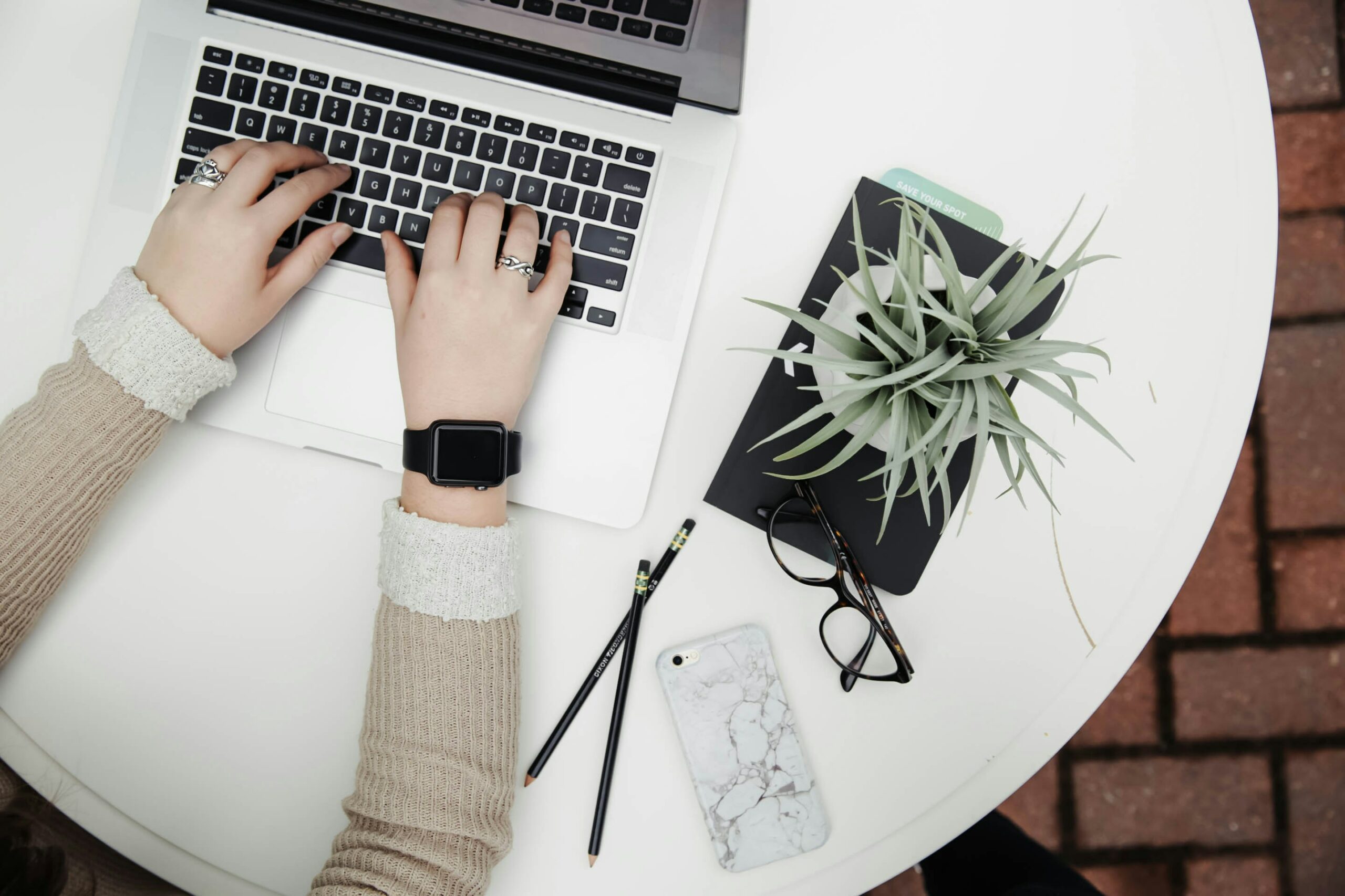 View of a woman's arms with beige sleeves and an apple watch, typing on a open laptop, on a white round table, with a spider plant and glasses to the right.